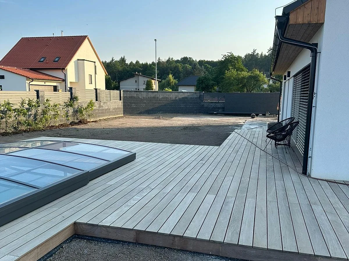Dřevěná terasa u domu se zastřešeným bazénem a dvěma křesly, okolo připravený terén